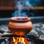 Uma panela de barro está em uma fogueira crepitante, com vapor subindo de dentro.