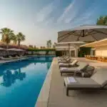 Área de lazer com piscina grande, cadeiras de descanso, guarda-sóis, plantas ao redor e um espaço externo com móveis confortáveis.