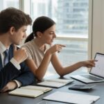 Dois jovens adultos sentados em uma mesa de escritório moderna, discutindo e olhando para um laptop com calculadora e calendário na mesa.
