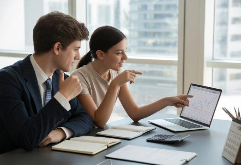 Dois jovens adultos sentados em uma mesa de escritório moderna, discutindo e olhando para um laptop com calculadora e calendário na mesa.