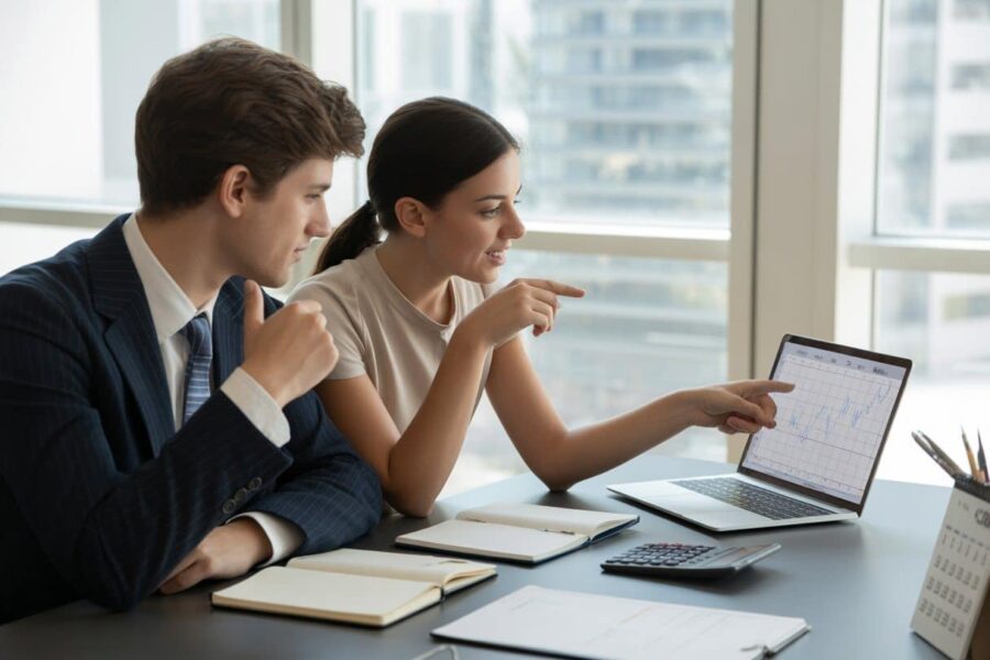Dois jovens adultos sentados em uma mesa de escritório moderna, discutindo e olhando para um laptop com calculadora e calendário na mesa.