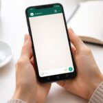 Pessoa segurando um smartphone com a tela do WhatsApp aberta, em uma mesa com itens de escritório ao fundo.