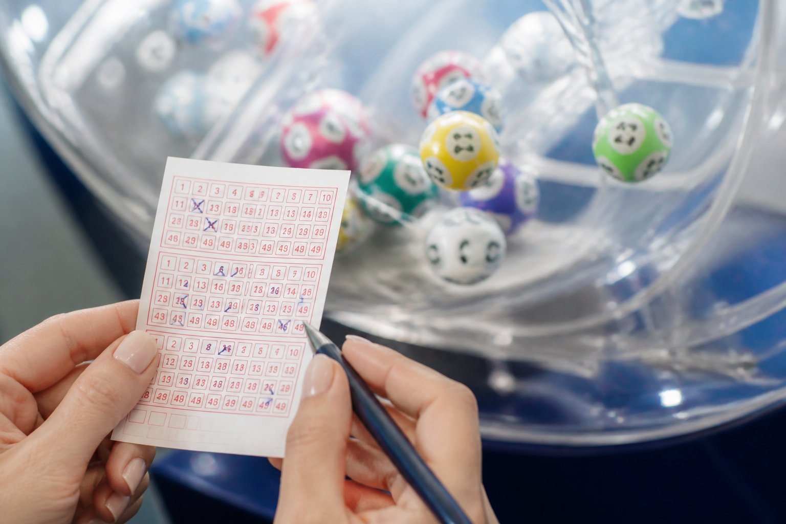 Mãos segurando um bilhete de loteria com números destacados, com uma máquina de sorteio e bolas numeradas ao fundo.